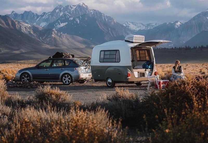 a small, fiberglass travel trailer being pulled by a Crossover SUV near the mountains