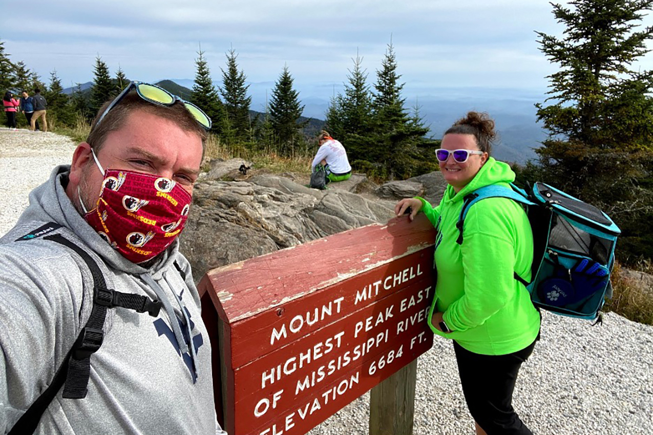 A couple standing in front of a sign and wearing a cat backpack.