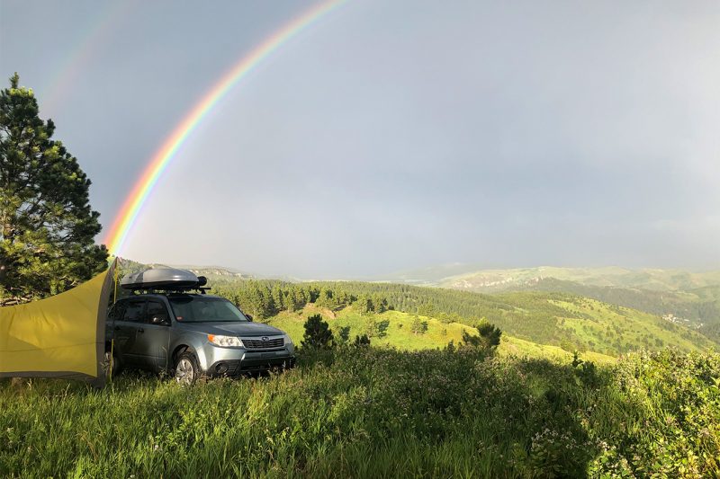 Car and tent set up near the field of a field with a rainbow in the sky.