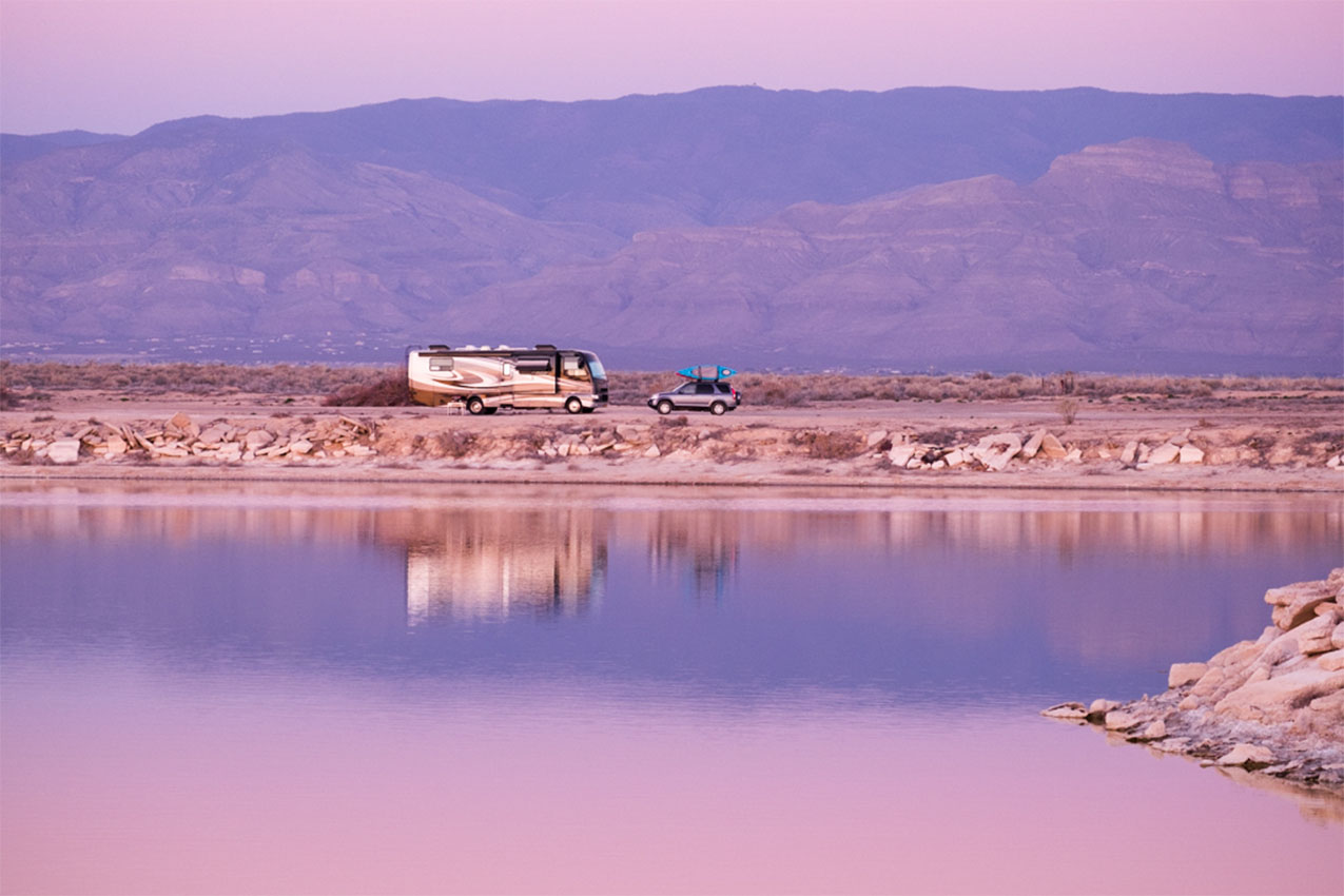 Pink sky and lake at sunset with class A and car parked on the shoreline.