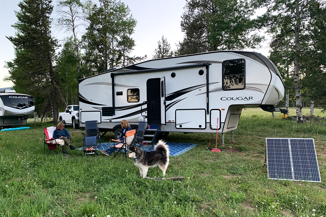 Two people and a dog sitting outside of two fifth-wheels.