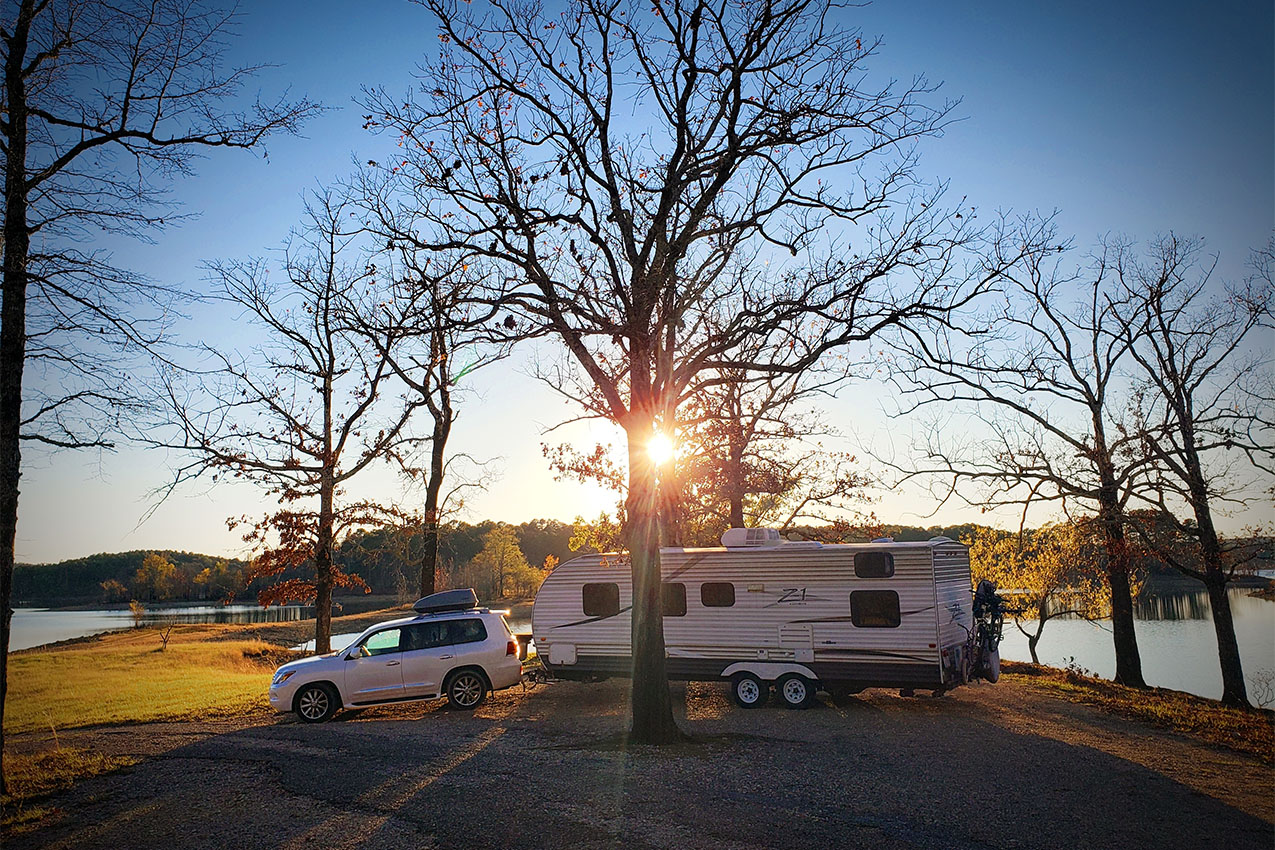 Truck and travel trailer parked under trees without leaves at sunset.