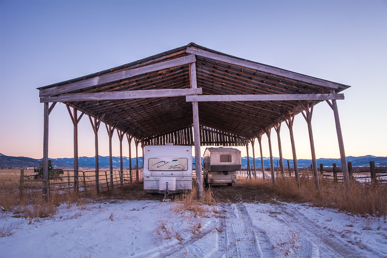 Two RVs stored under a hay barn with snow on the ground.