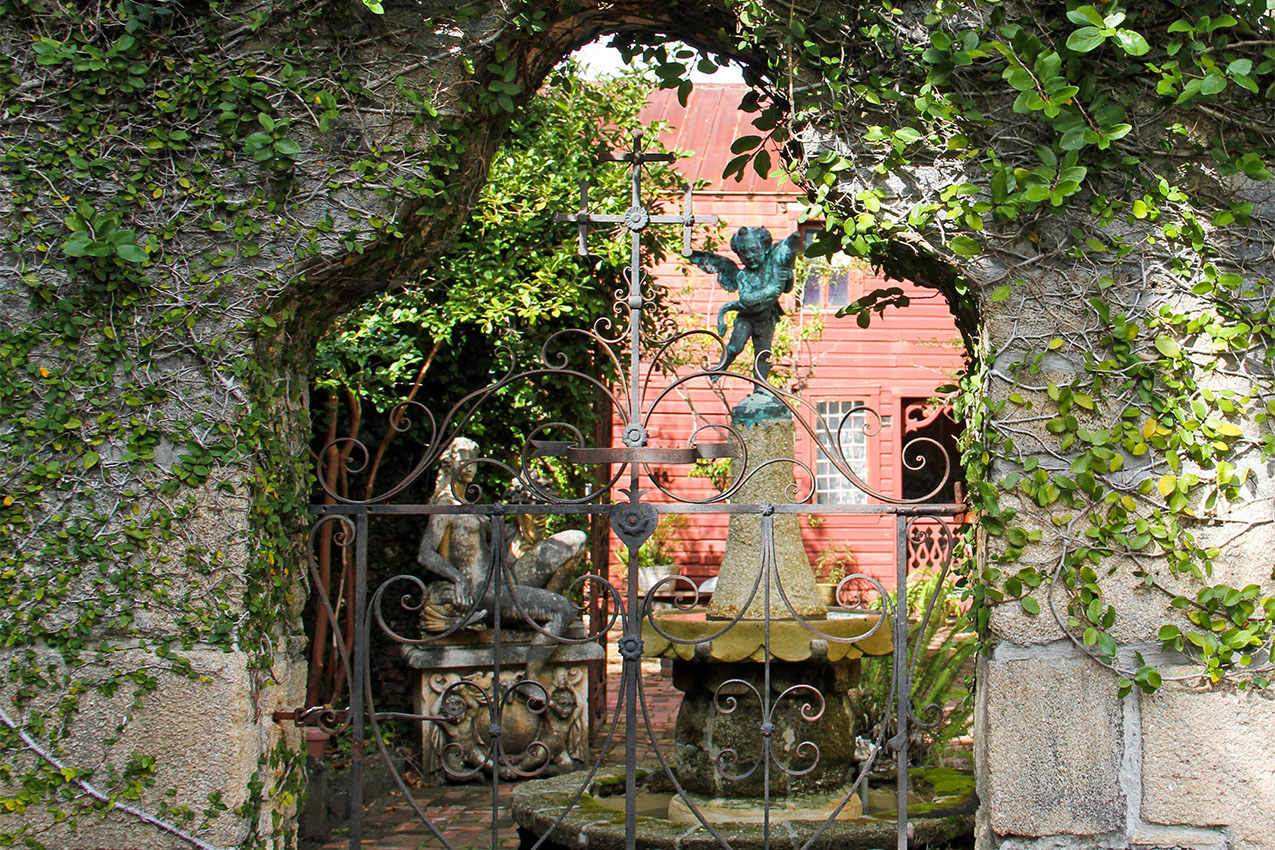 Ivy covered stone wall looking through iron gate into courtyard with statues.