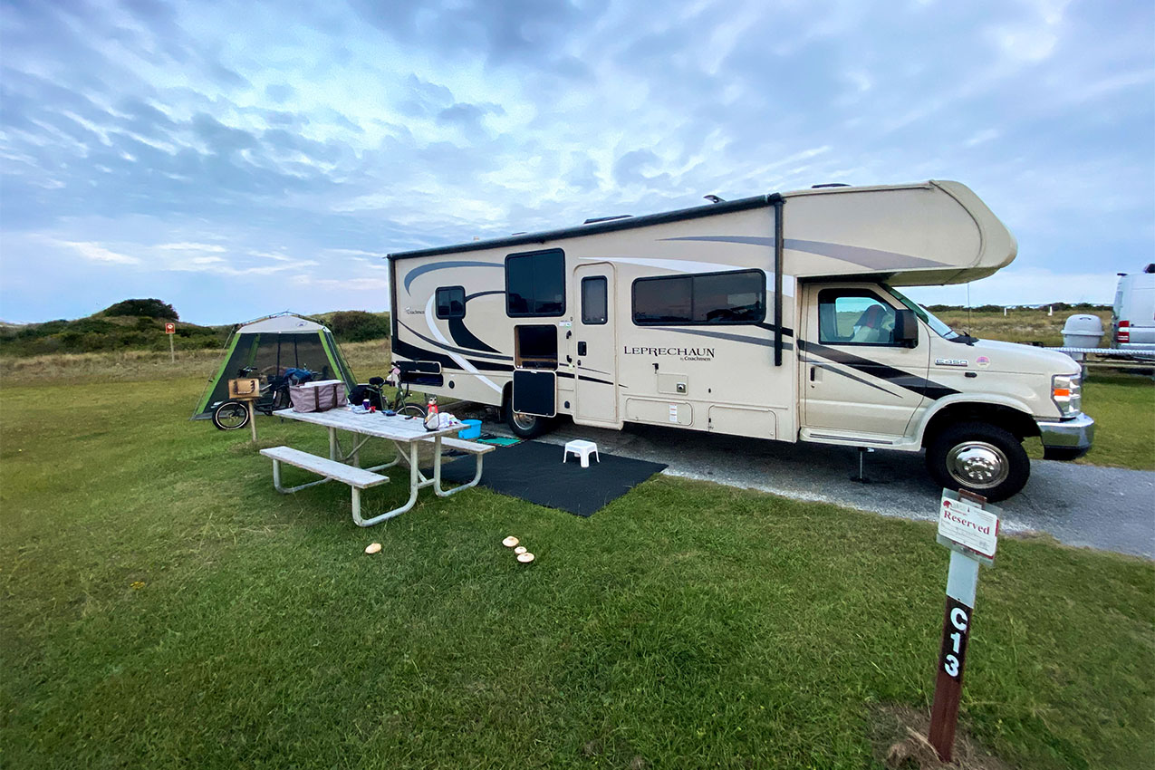 Large class C RV parked in front of a picnic table under fluffy blue clouds.