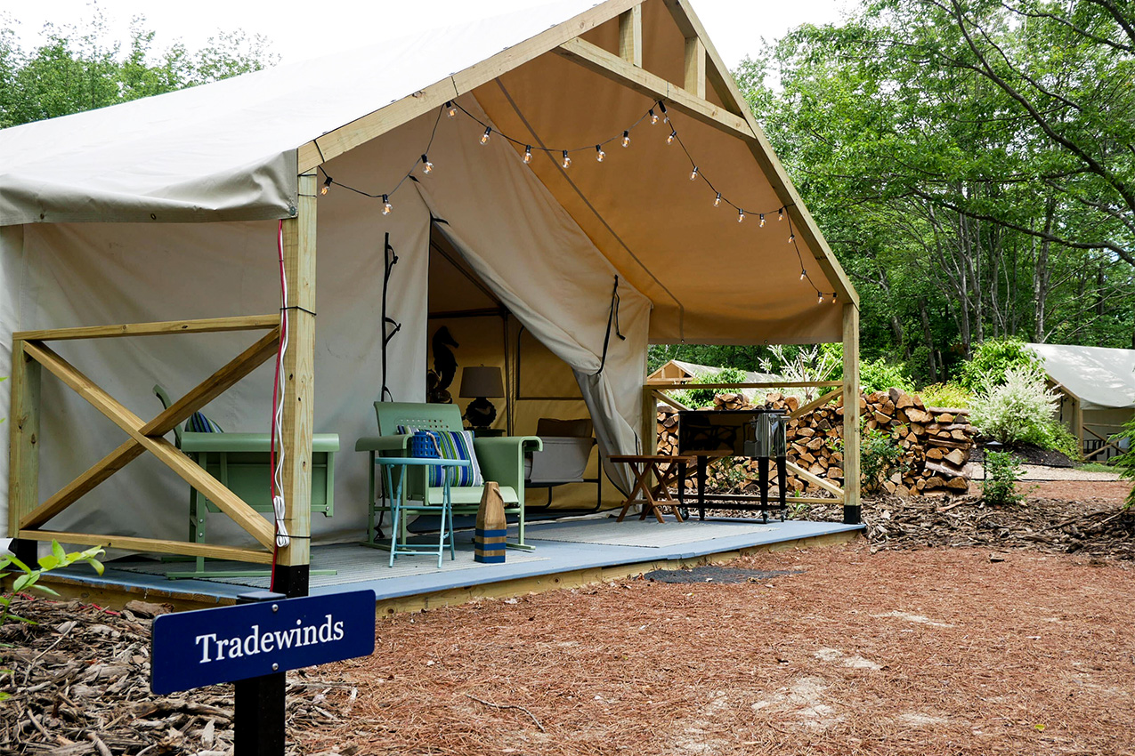 Canvas tent in the woods with camp chairs on the porch and firewood.