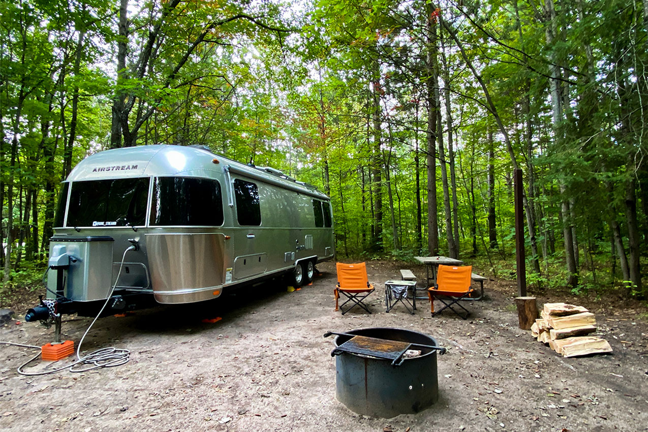 Airstream parked in a spot next to camp chairs, fire ring, and stack of wood.