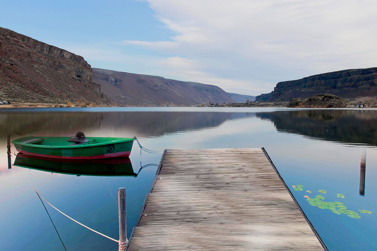 Wood dock leading to a green boat over blue lake and mountains in the background.
