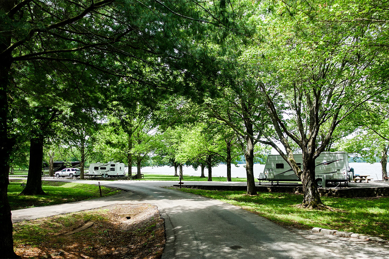 Two RVs parked in a campground under trees next to a lake.