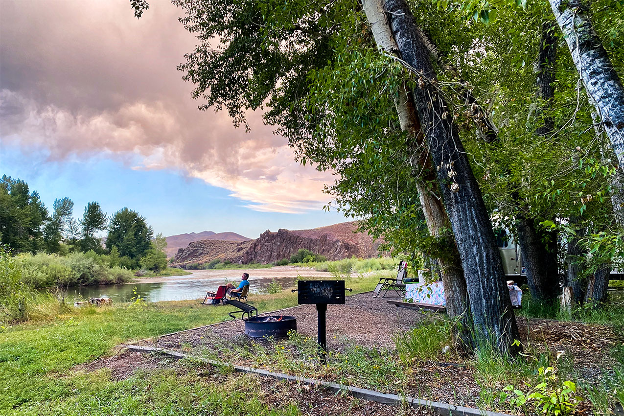 Man sitting in a camp chair by fire ring under pink clouds.