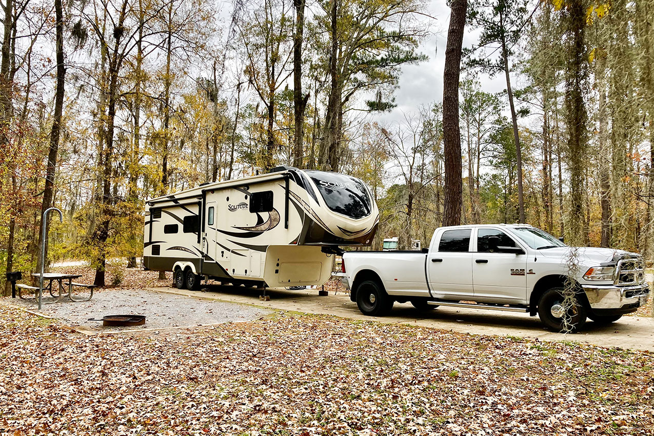 Fifth wheel and truck parked in a campsite under tall trees.