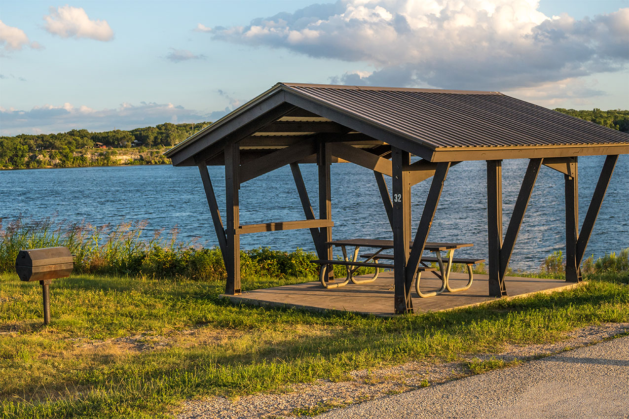 Covered picnic table and grill in front of a lake during golden hour.