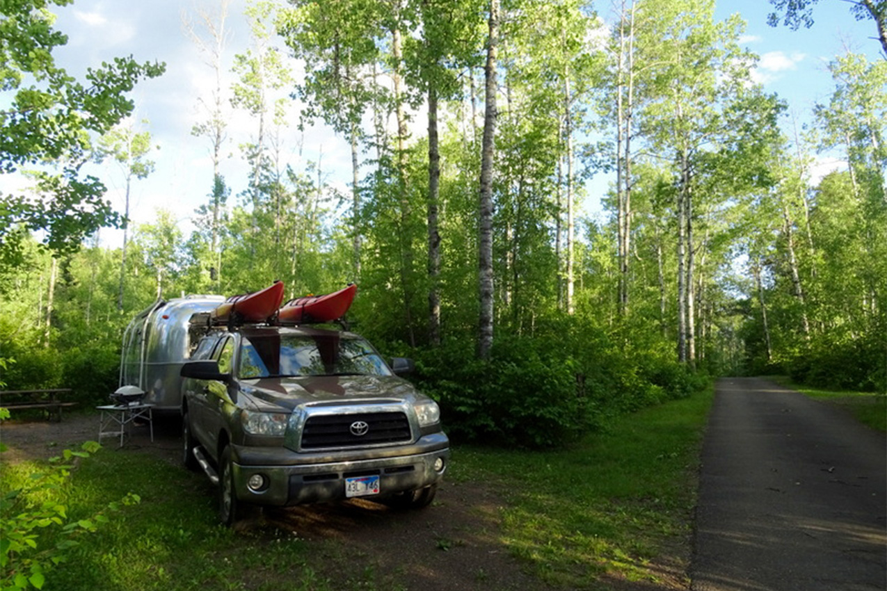 Truck with kayaks on roof attached to an Airstream in a campsite.