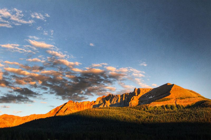 Tops of mountains lit up in sunset with cloud hovering aboe.