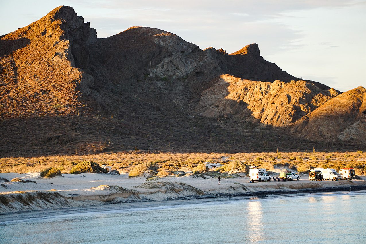 Several RVs parked along the ocean with a big mountain in the background.
