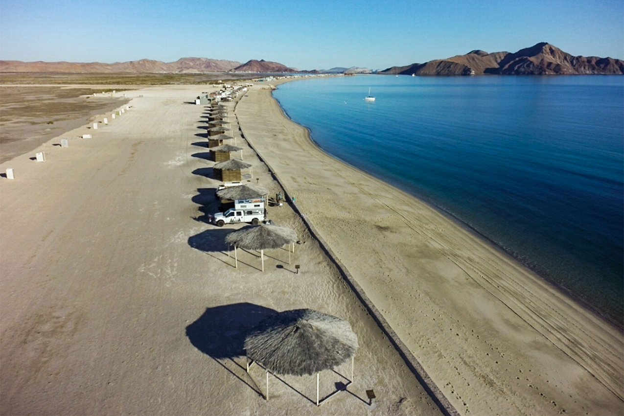 Aerial view of sandy beach lined with several palapas and a truck camper.