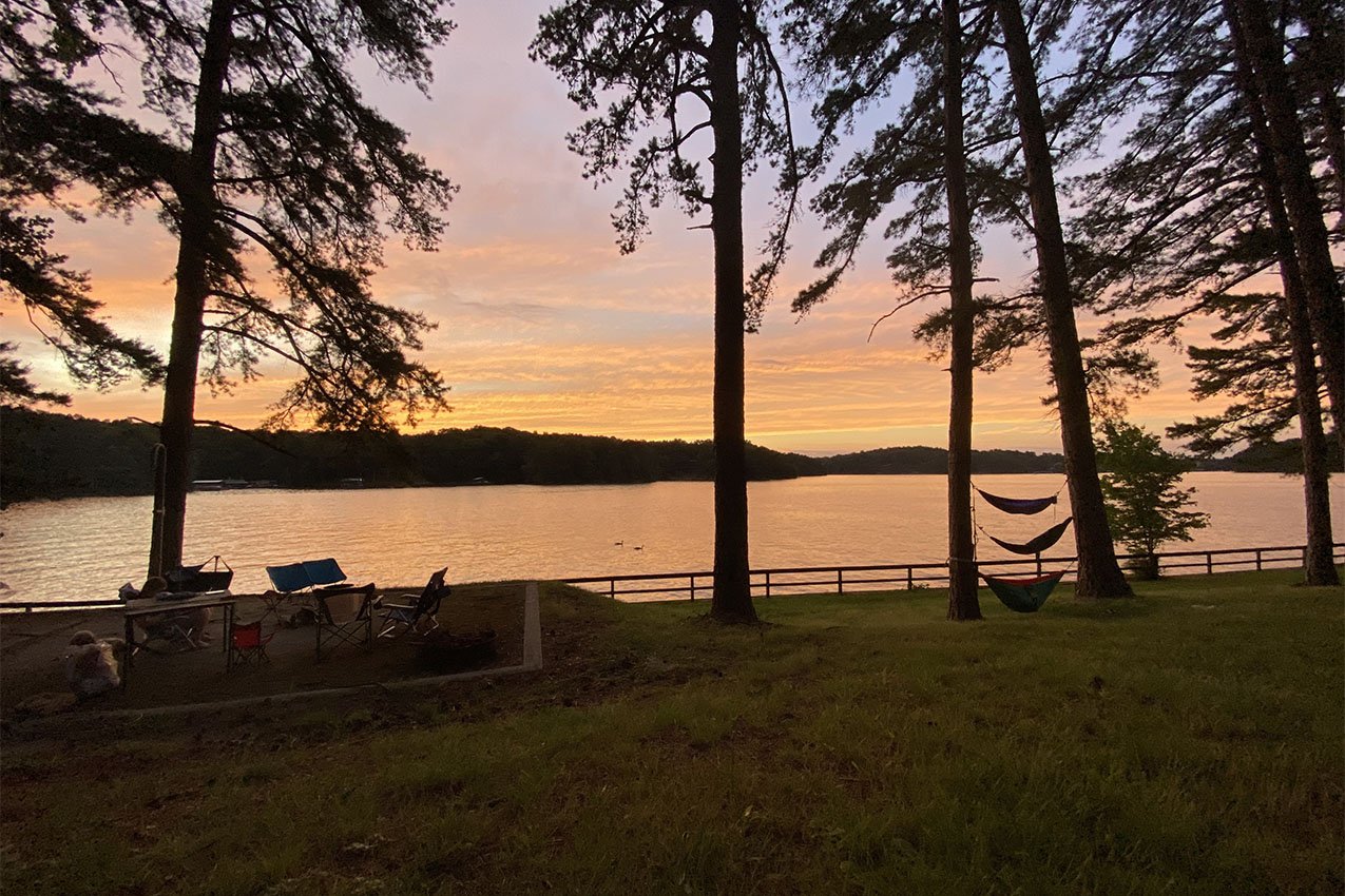 Silhouette of hammocks hanging between trees and camp chairs next to a lake during sunset.