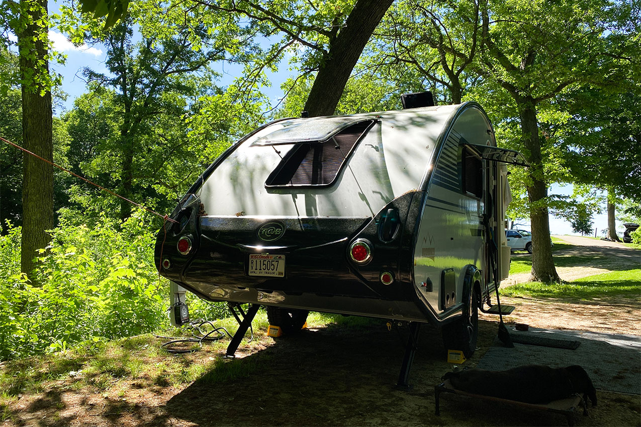 Teardrop camper parked in a heavily wooded campsite with a dog sleeping outside.