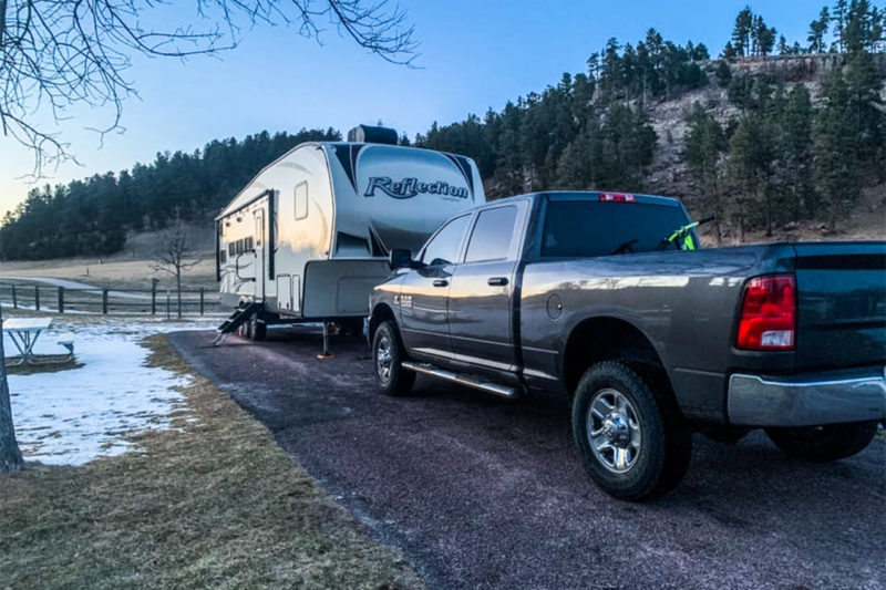 Truck parked in front of a fifth wheel under a tree with some snow on the ground.