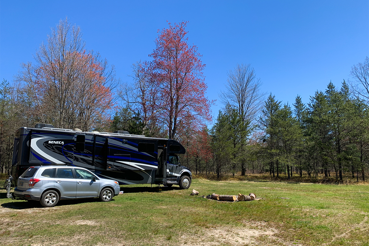 Large class C and car parked next to a fire ring in a field with fall colored trees.