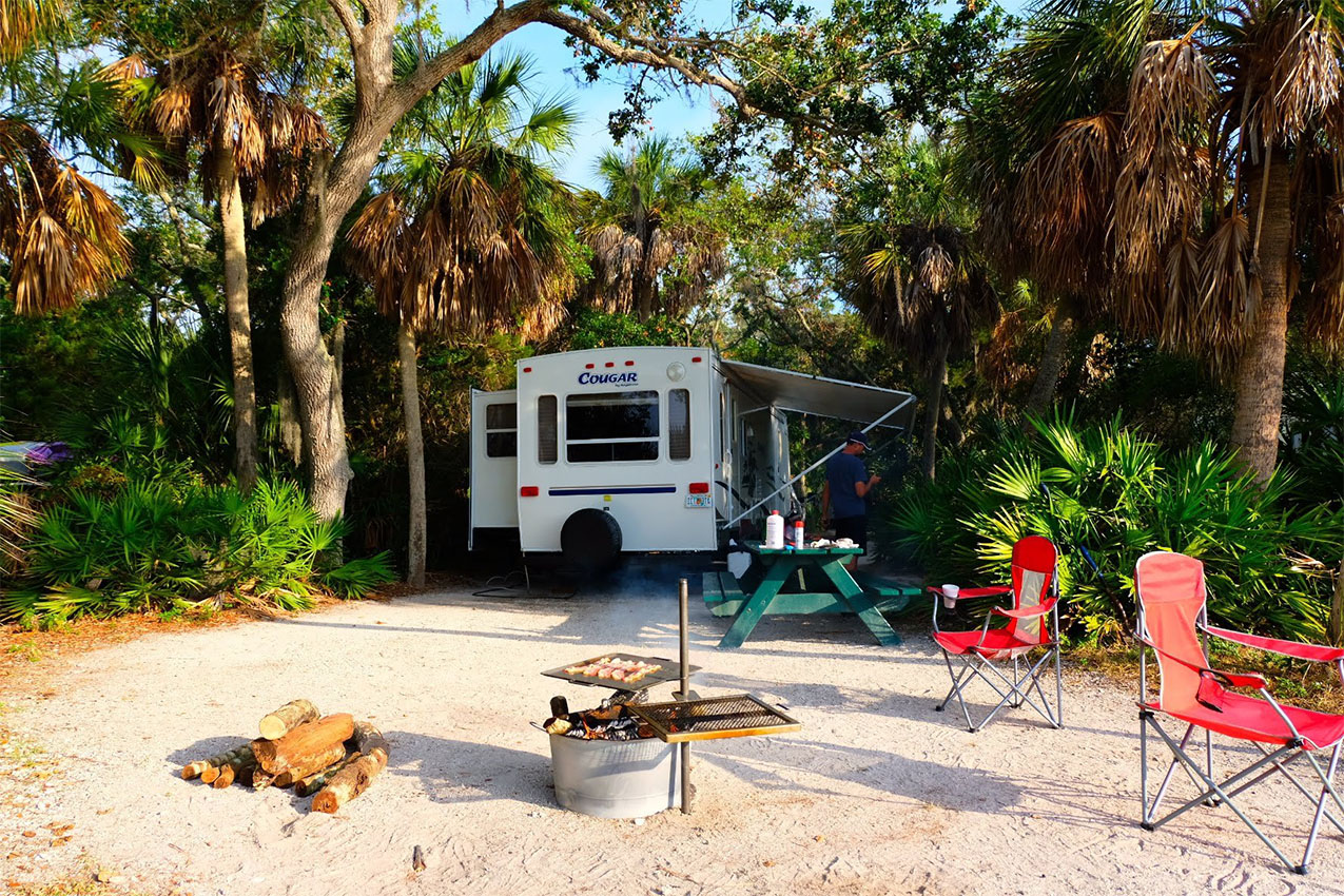 Camp chairs and campfire set up on a sandy area by an RV.