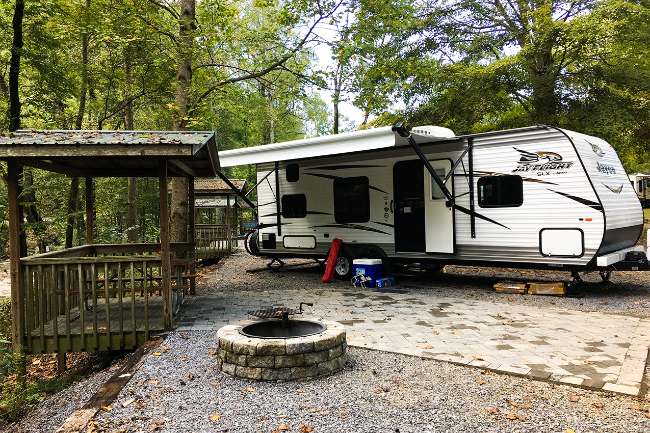 Travel trailer parked under trees next to a wooden picnic shelter and stone fire ring.