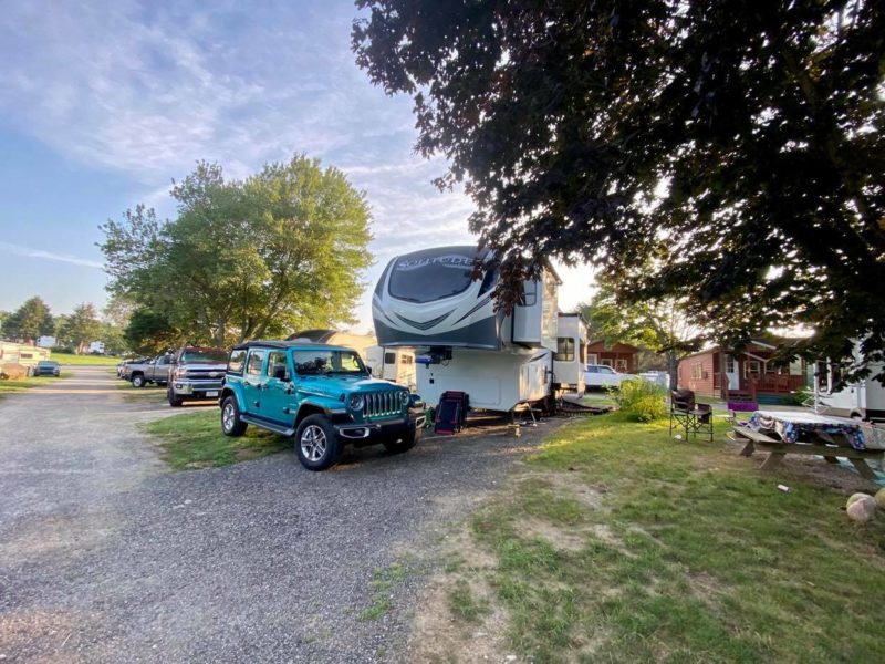 Jeep parked in front of a large fifth-wheel in a gravel RV site next to a big tree.