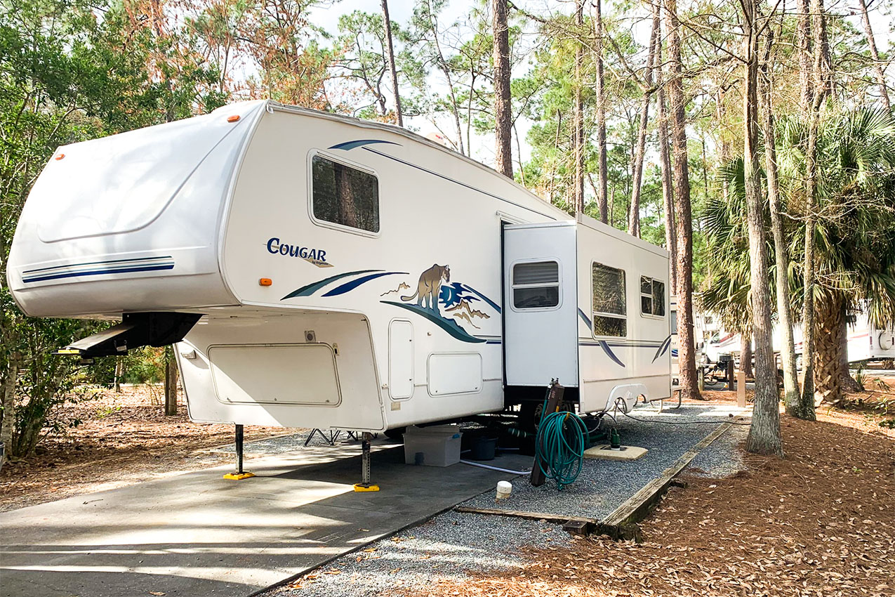 Large white fifth-wheel parked on a concrete pad in the woods of an RV park.