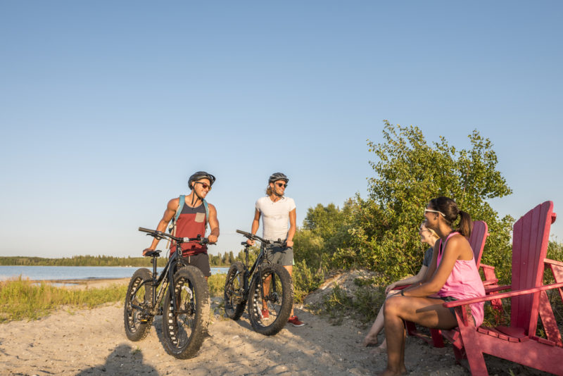 two people ride bikes in the sand and two people sit in red chairs under a clear blue sky