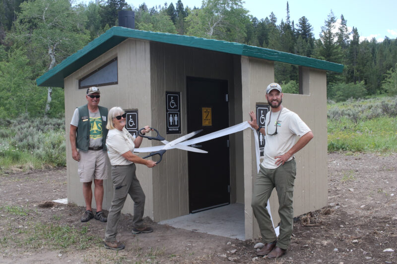 Three people standing next to a vault toilet, one of them cutting a ribbon with oversized scissors