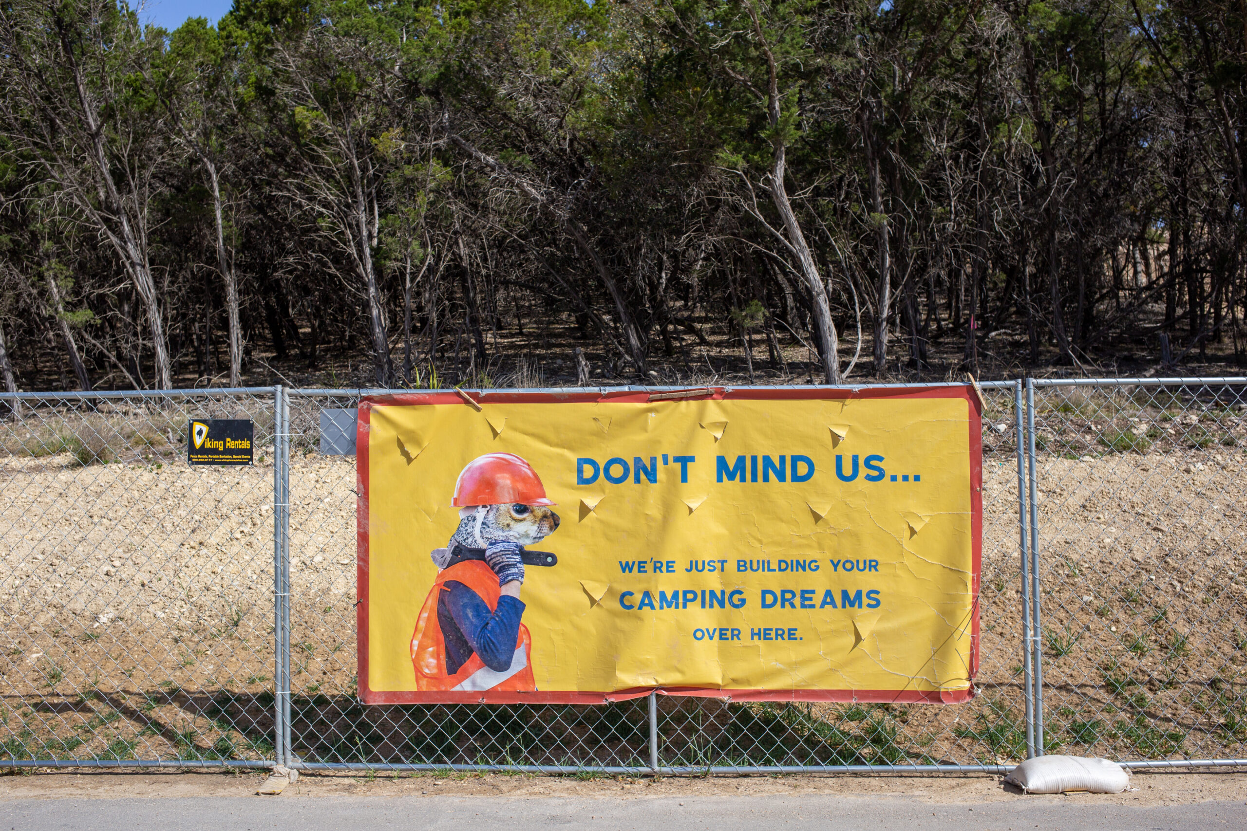 a fence and a sign that says "don't mind us we're just building your camping dreams over here"