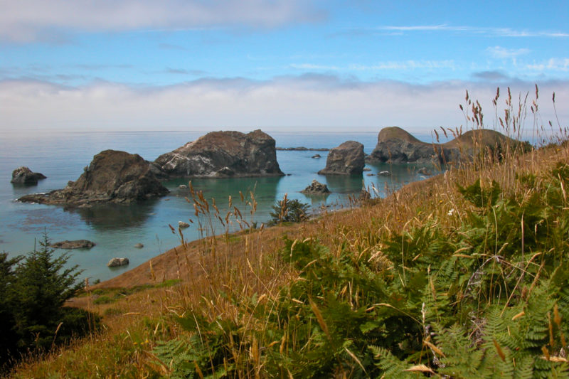 a shoreline with rocks jutting out of the water under a blue sky and low gray clouds