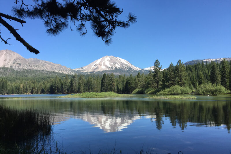 a snowy mountain peak is reflected in a clear lake under clear blue skies surrounded by pine trees