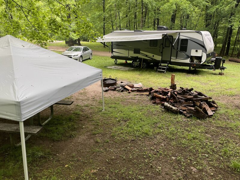 a campsite in the woods with an RV and car parked next to a wood pile and white pop up tent