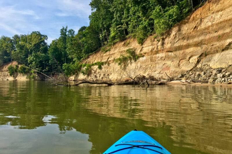 the tip of a blue kayak going through the water with rocks and trees on the shoreline