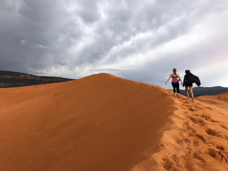 two people walk on a large sand dune under cloudy skies