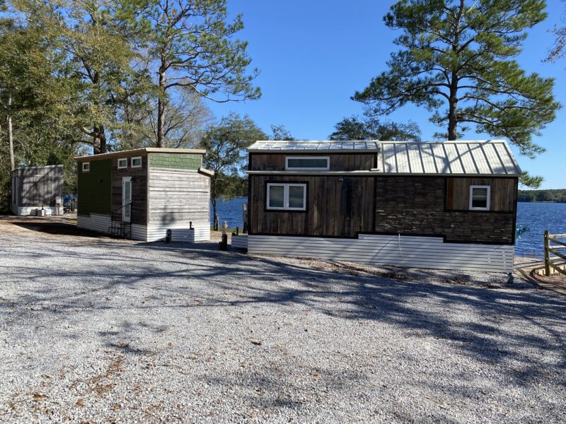 two tiny homes along the water's edge at a gravel campsite surrounded by trees