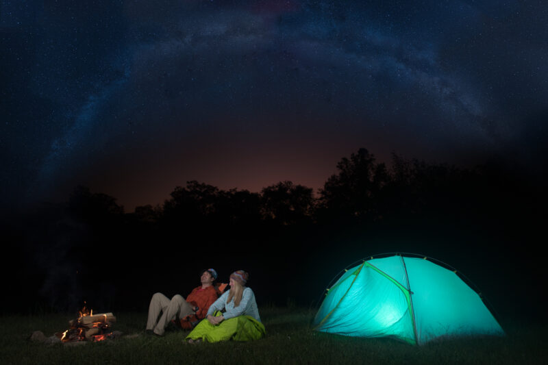 Campers sit by an illuminated tent as the night sky comes alive with a dazzling array of stars.
