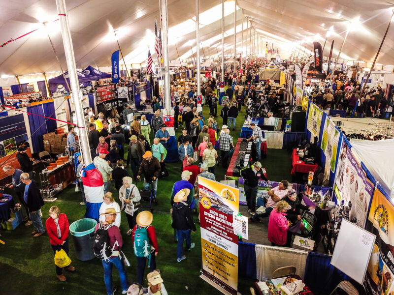 A view looking down the long length of a white tent lined with exhibitor booths of various sizes and configurations and packed with people looking at the booth offerings.