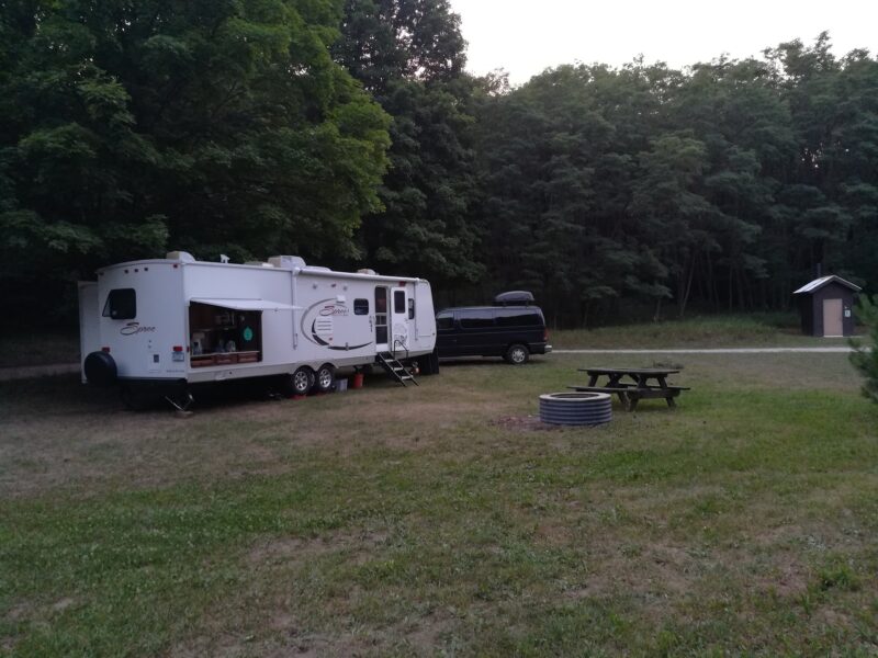 Large travel trailer parked near an open campsite with a forest in the backdrop