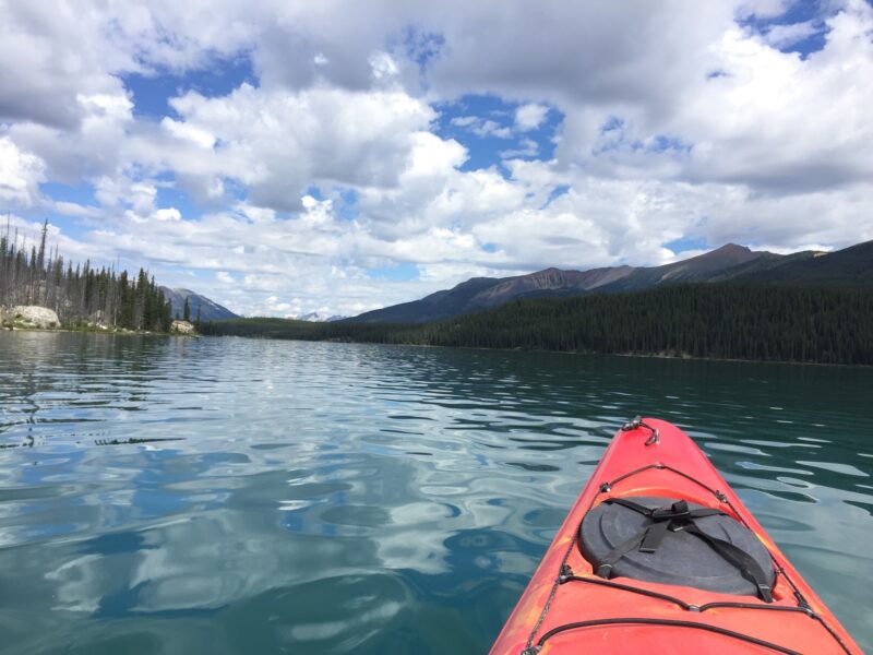 the tip of an orange red kayak in a blue lake with mountains in the background under a blue sky with fluffy white clouds
